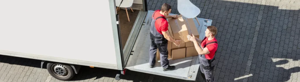 Local movers in Houston wearing red uniforms carefully loading cardboard boxes onto a moving truck’s lift gate on a sunny driveway