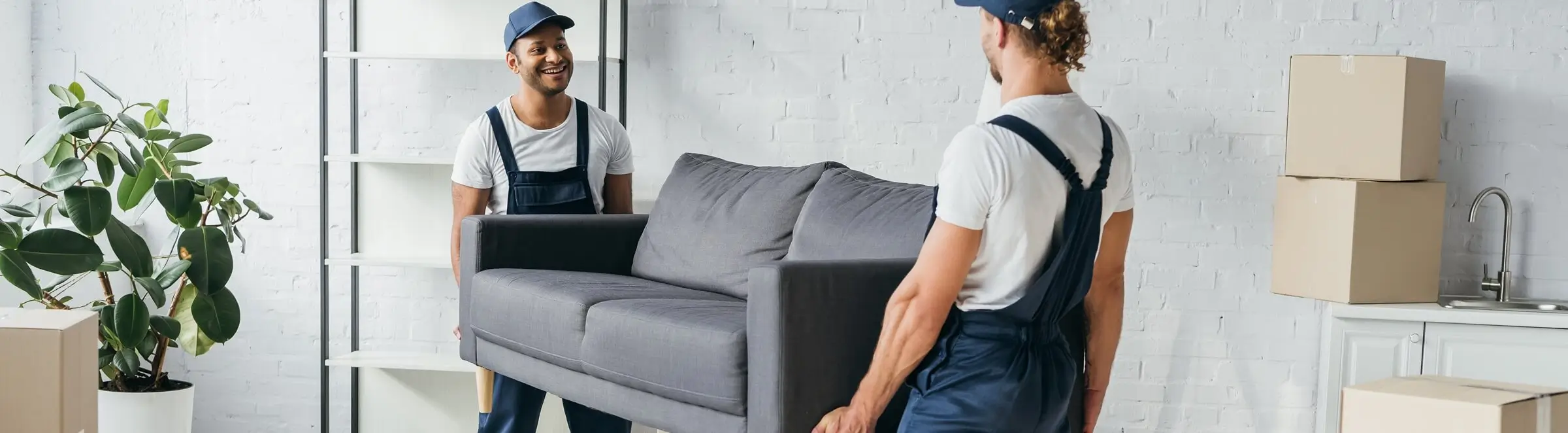 Two movers in uniforms lifting a gray sofa inside a bright room with moving boxes and a potted plant.